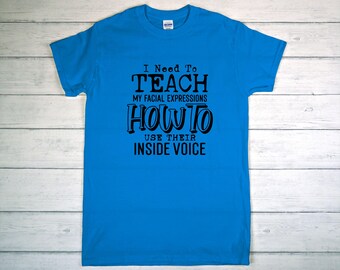 May include: A bright blue t-shirt with the text "I Need To Teach My Facial Expressions How To Use Their Inside Voice" in black. The shirt is made of a soft material and has a crew neck.