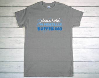 May include: A gray t-shirt with the text "please hold I'M MENTALLY BUFFERING" in white and blue. The shirt is made of a soft material and has short sleeves. The text is centered on the front of the shirt.