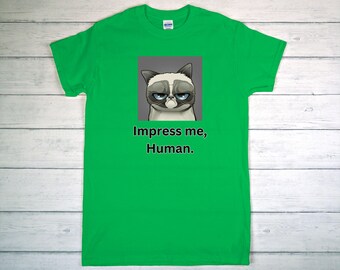 May include: Bright green t-shirt featuring a grumpy cat graphic with the text "Impress me, Human." The cat has a gray and white face with blue eyes. The shirt is a short-sleeved crew neck.