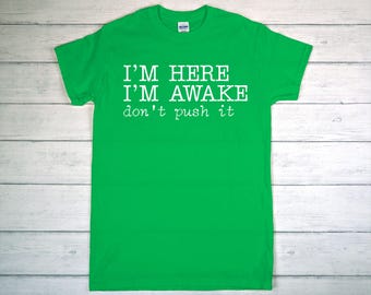 May include: A bright green t-shirt with the text "I'M HERE I'M AWAKE don't push it" in white. The shirt is made of a soft material and has short sleeves. The text is centered on the front of the shirt.