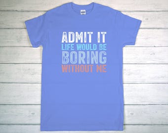 May include: A light blue t-shirt with the text "ADMIT IT LIFE WOULD BE BORING WITHOUT ME" in white, light blue, and coral lettering. The shirt is displayed on a light wood surface.