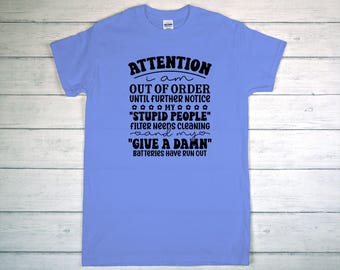 May include: A light blue t-shirt with black text. The text reads "ATTENTION I am OUT OF ORDER UNTIL FURTHER NOTICE MY 'STUPID PEOPLE' FILTER NEEDS CLEANING and my 'GIVE A DAMN' BATTERIES HAVE RUN OUT."
