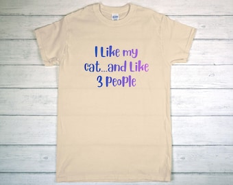 May include: A cream-colored t-shirt with the text "I Like my cat...and Like 3 People" in blue and purple. The shirt is made of a soft material and has a crew neck.