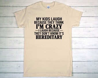 May include: A light beige t-shirt with the text "MY KIDS LAUGH BECAUSE THEY THINK I'M CRAZY - I LAUGH BECAUSE THEY DON'T KNOW IT'S HEREDITARY" in black block letters. The shirt is displayed on a light wood-grain surface.