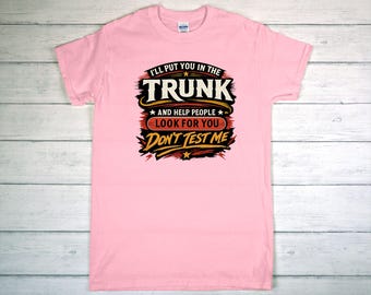 May include: A light pink t-shirt with the text "I'LL PUT YOU IN THE TRUNK and help people LOOK FOR YOU DON'T TEST ME" in a black, red, and yellow design. The shirt is a crew neck style.