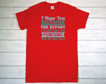 May include: A red t-shirt with the text "I Hope You APPRECIATE THE EFFORT I've put into not PUNCHING You In The Face" in light blue. The shirt is made of a soft material and has short sleeves.