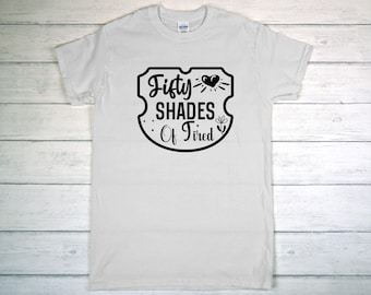 May include: White t-shirt with a black graphic that reads "Fifty Shades Of Tired" inside a shield design. The design includes a heart with rays and a small flower. The shirt is displayed on a light wood-grain surface.