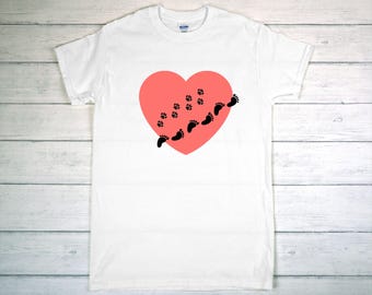 May include: White t-shirt featuring a coral-colored heart with black paw prints and footprints. The design suggests a theme of pets or family, with a simple and clean aesthetic. The shirt is made of a soft material.