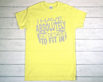 May include: A yellow t-shirt with the text "I HAVE ABSOLUTELY NO DESIRE TO FIT IN" in gray lettering. The shirt is made of a soft material and has short sleeves and a crew neck.