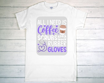 May include: White t-shirt with a graphic design. The design features the text "ALL I NEED IS Coffee SCRUBS AND RUBBER GLOVES" in purple and white, along with a coffee cup and a stethoscope. The shirt is displayed on a light wood background.