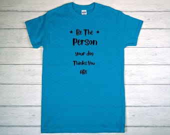 May include: A turquoise t-shirt with the text "Be The Person your dog Thinks You ARE" in black. The shirt has a crew neck and short sleeves. The text is centered on the front of the shirt.