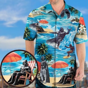 Op de afbeelding: Een shirt met korte mouwen met een tropisch strandontwerp met Darth Vader die surft en ontspant. Het shirt is overwegend blauw met illustraties van palmbomen, golven en parasols. Het ontwerp bevat een cirkelvormige inzet met een strandscène.