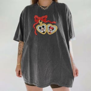 Op de afbeelding: Donkergrijs T-shirt met een Mickey en Minnie Mouse ontwerp. De afbeelding toont Mickey en Minnie in hartvormige kaders, verbonden door een rode strik. Het shirt heeft korte mouwen en een relaxte pasvorm, geschikt voor casual wear.