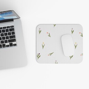 May include: A white computer mouse sits on a white mouse mat with a floral pattern. The pattern features small green stems with pink, yellow, and purple flowers.