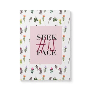 May include: A white notebook with a pink rectangle in the center. The rectangle has the text "SEEK #HIS FACE" in black and red lettering. The notebook is covered in a pattern of small, colorful flowers.