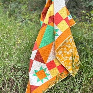 Op de afbeelding: Een kleurrijke quilt met geometrische patronen en een bloemenprint op de achterkant. De quilt heeft oranje, gele, groene en witte blokken en vormen. De quilt hangt aan een tak in een grasrijk gebied.