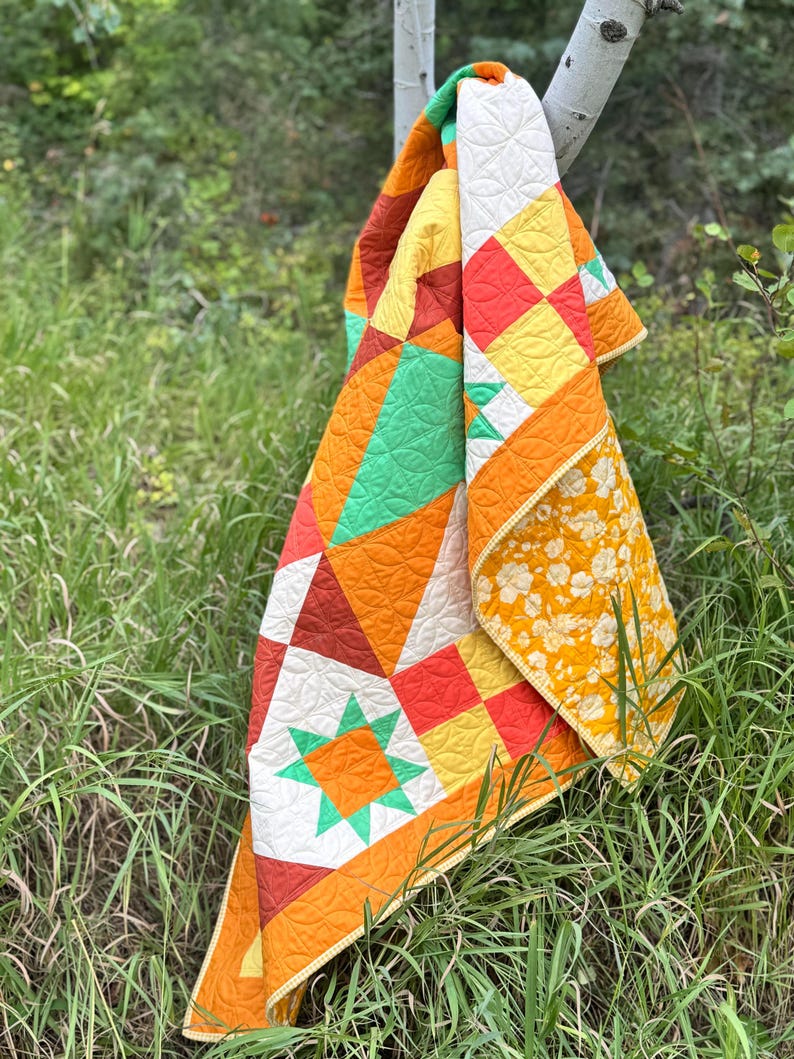 Op de afbeelding: Een kleurrijke quilt met geometrische patronen en een bloemenprint op de achterkant. De quilt heeft oranje, gele, groene en witte blokken en vormen. De quilt hangt aan een tak in een grasrijk gebied.