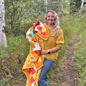 Op de afbeelding: Een kleurrijke quilt met een patchwork-ontwerp met vierkanten in oranje, geel, rood en groen. De quilt heeft een bloemenrand en wordt vastgehouden door een persoon in een gele top en een blauwe spijkerbroek.