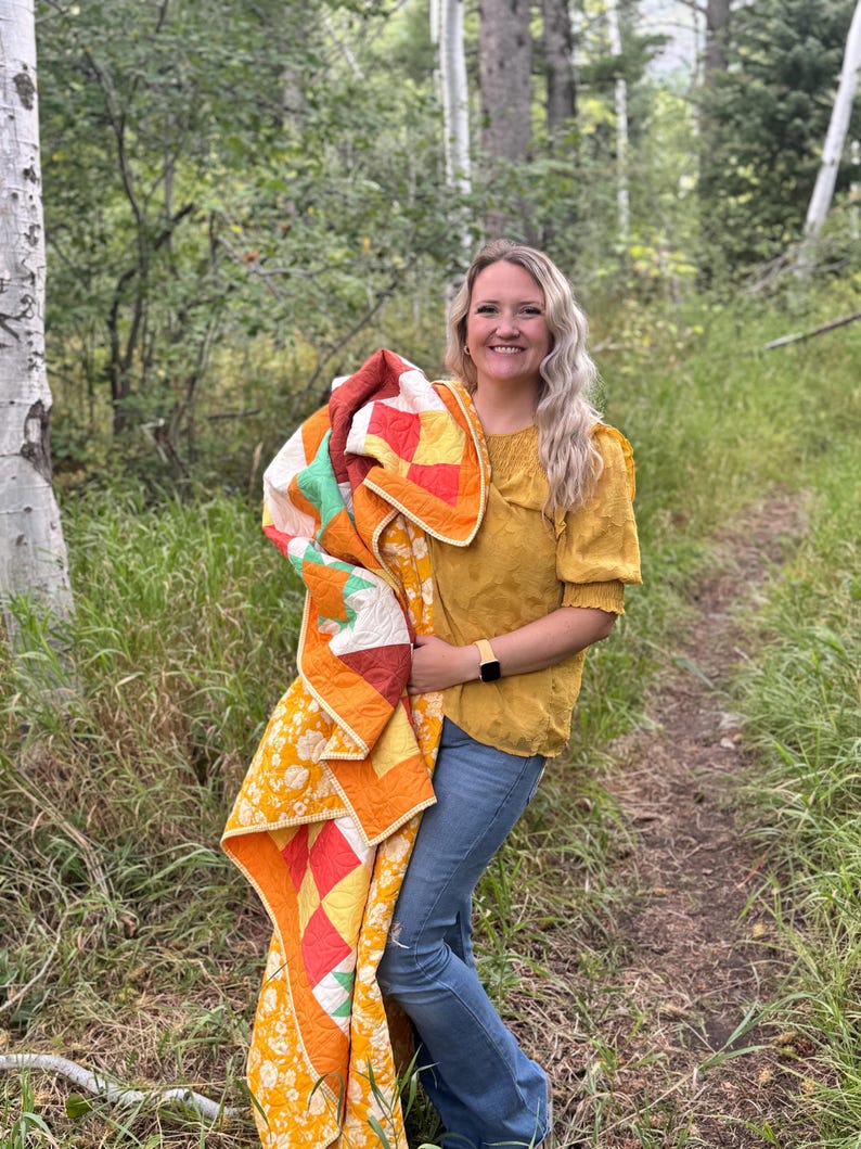 Op de afbeelding: Een kleurrijke quilt met een patchwork-ontwerp met vierkanten in oranje, geel, rood en groen. De quilt heeft een bloemenrand en wordt vastgehouden door een persoon in een gele top en een blauwe spijkerbroek.