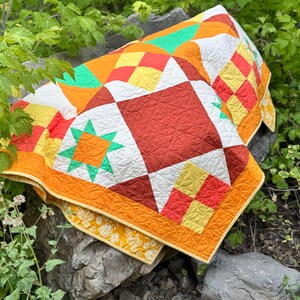 Op de afbeelding: Een kleurrijke, handgemaakte quilt met een geometrisch patroon. De quilt heeft vierkanten en ruiten in tinten oranje, rood, geel, wit en groen. De quilt wordt buiten op een rots getoond en toont het levendige ontwerp.