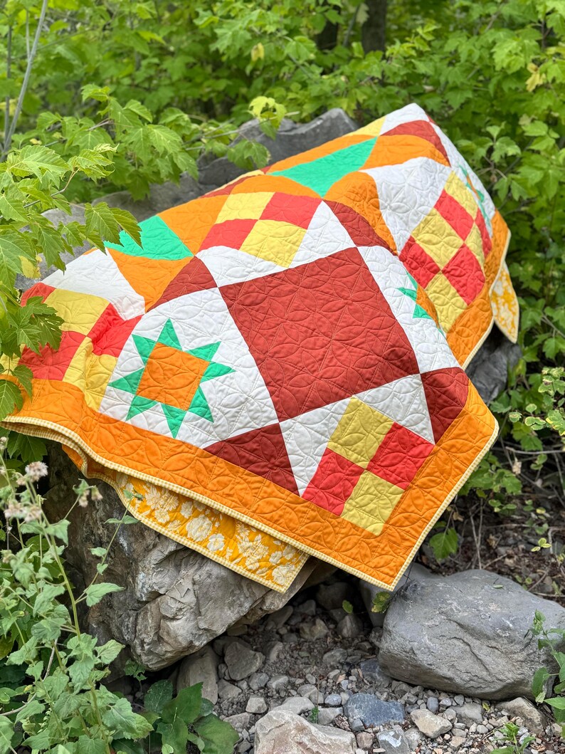 Op de afbeelding: Een kleurrijke, handgemaakte quilt met een geometrisch patroon. De quilt heeft vierkanten en ruiten in tinten oranje, rood, geel, wit en groen. De quilt wordt buiten op een rots getoond en toont het levendige ontwerp.