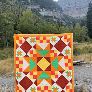 Op de afbeelding: Een kleurrijke quilt met een geometrisch sterpatroon. De quilt heeft een levendige mix van oranje, gele, groene en rode vierkanten, ruiten en sterren. De quilt heeft een effen oranje rand en wordt buiten getoond.