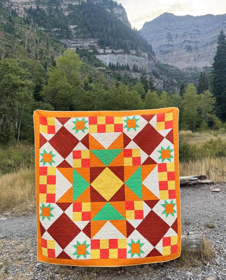 Op de afbeelding: Een kleurrijke quilt met een geometrisch sterpatroon. De quilt heeft een levendige mix van oranje, gele, groene en rode vierkanten, ruiten en sterren. De quilt heeft een effen oranje rand en wordt buiten getoond.