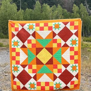 Op de afbeelding: Een kleurrijk quilt met een geometrisch sterrenpatroon. De quilt heeft een centrale gele ruit, omringd door oranje, groene en rode vormen. De rand is effen oranje. De quilt wordt buiten getoond.