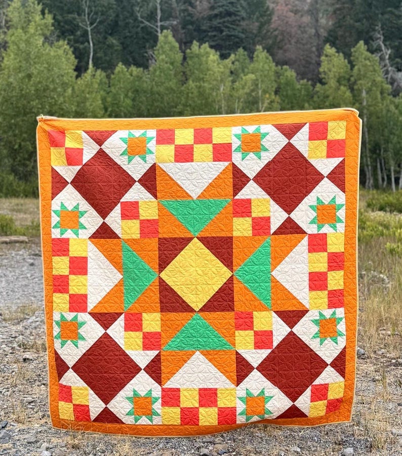 Op de afbeelding: Een kleurrijk quilt met een geometrisch sterrenpatroon. De quilt heeft een centrale gele ruit, omringd door oranje, groene en rode vormen. De rand is effen oranje. De quilt wordt buiten getoond.
