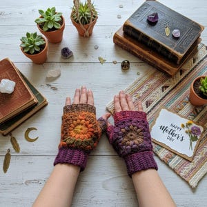 Puede incluir: Un par de guantes sin dedos de ganchillo hechos a mano en tonos morados, naranjas y marrones. Los guantes presentan un diseño floral en el dorso de la mano. También se ve una tarjeta de "Feliz Día de la Madre".