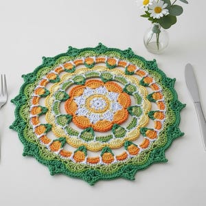 May include: A colorful, crocheted placemat with a floral design. The circular mat features concentric rings of green, yellow, orange, and white. The outer edge has a scalloped border. A fork, knife, and small vase of flowers are in the background.