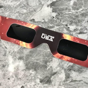 Pode incluir: Um par de óculos de eclipse preto com um design vermelho, laranja e preto que representa um eclipse solar. Os óculos têm o texto "ECLIPSE USA" impresso neles.