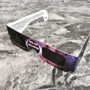 May include: A pair of cardboard solar eclipse glasses with a black and white galaxy print. The glasses have the words "Solar Eclipse" printed on them.