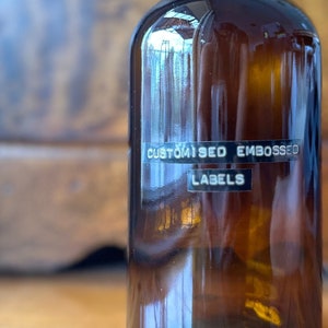 Puede incluir: Una botella de vidrio marrón con una tapa negra y una etiqueta negra que dice "CUSTOMISED EMBOSSED LABELS".