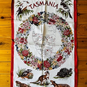 Puede incluir: Una toalla de cocina blanca con un borde rojo presenta un mapa de Tasmania, Australia, rodeado de una corona de flores nativas. El mapa incluye las principales ciudades y pueblos. Debajo del mapa hay ilustraciones de animales nativos, incluyendo un demonio de Tasmania, un canguro, un equidna, un ornitorrinco y un tigre de Tasmania. El texto "TASMANIA" est&aacute; impreso en rojo en la parte superior de la toalla.