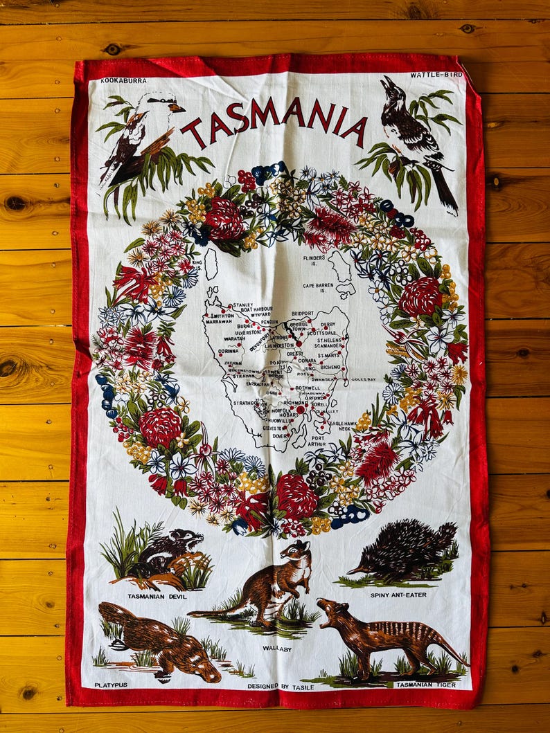 Puede incluir: Una toalla de cocina blanca con un borde rojo presenta un mapa de Tasmania, Australia, rodeado de una corona de flores nativas. El mapa incluye las principales ciudades y pueblos. Debajo del mapa hay ilustraciones de animales nativos, incluyendo un demonio de Tasmania, un canguro, un equidna, un ornitorrinco y un tigre de Tasmania. El texto "TASMANIA" est&aacute; impreso en rojo en la parte superior de la toalla.