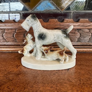 May include: A porcelain figurine of a white and brown terrier dog with two smaller terrier puppies. The figurine is on a white base.