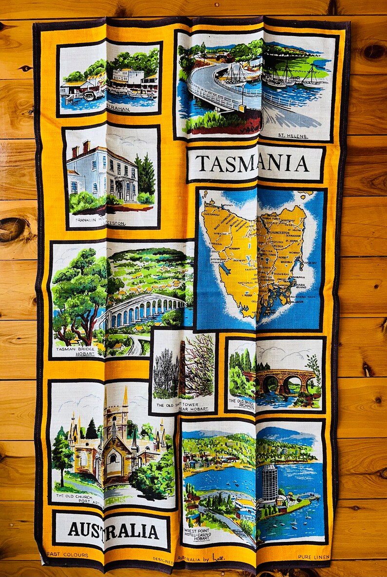 Puede incluir: Una toalla de t&eacute; amarilla con un mapa de Tasmania, Australia. La toalla presenta im&aacute;genes de varios puntos de referencia, incluidos puentes, iglesias y edificios. El texto "TASMANIA" y "AUSTRALIA" est&aacute; impreso en la toalla.