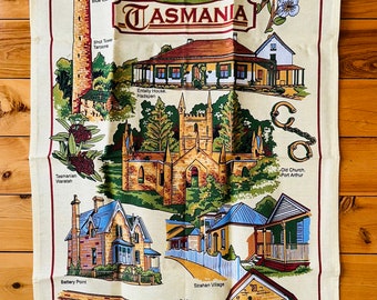 Op de afbeelding: Een vintage theedoek met illustraties van historische bezienswaardigheden in Tasmanië, Australië. De theedoek is crèmekleurig met rode, bruine en groene accenten. De tekst "Historic Tasmania" is gedrukt in een rode banner bovenaan de theedoek.