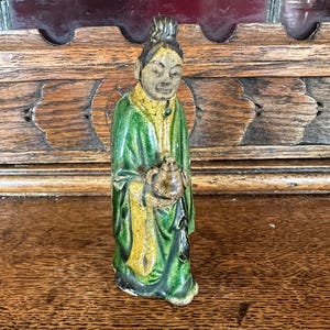 Puede incluir: Una figurita de cerámica esmaltada en verde y amarillo que representa a una persona sosteniendo una pequeña olla. La figurita está de pie sobre una superficie de madera.