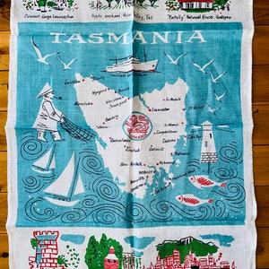 Puede incluir: Una toalla de t&eacute; azul y blanca con un mapa de Tasmania, Australia. El mapa muestra la ubicaci&oacute;n de varias ciudades y pueblos, incluyendo Hobart, Launceston y Burnie. La toalla de t&eacute; tambi&eacute;n presenta ilustraciones de un faro, un barco y un pescador. El texto "TASMANIA" est&aacute; impreso en letras grandes en la parte superior de la toalla.