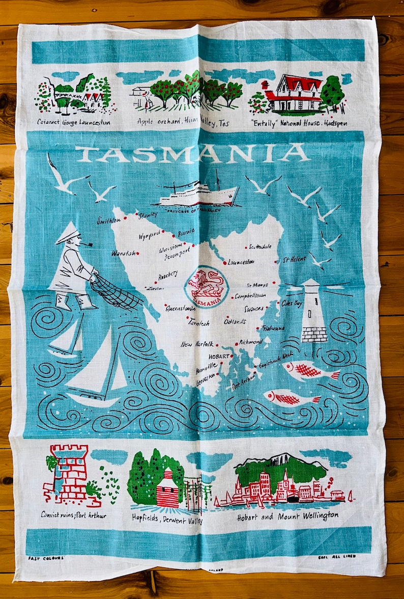 Puede incluir: Una toalla de t&eacute; azul y blanca con un mapa de Tasmania, Australia. El mapa muestra la ubicaci&oacute;n de varias ciudades y pueblos, incluyendo Hobart, Launceston y Burnie. La toalla de t&eacute; tambi&eacute;n presenta ilustraciones de un faro, un barco y un pescador. El texto "TASMANIA" est&aacute; impreso en letras grandes en la parte superior de la toalla.