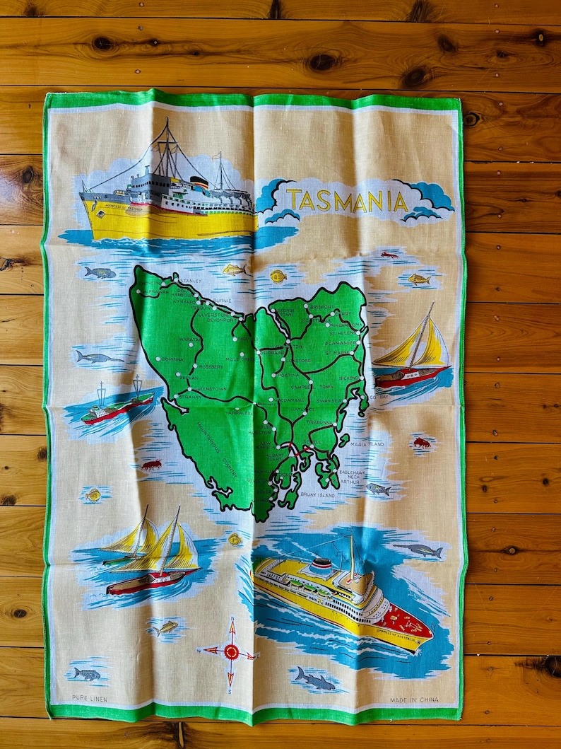 Puede incluir: Un pa&ntilde;o de cocina vintage con un mapa de Tasmania, Australia. El mapa es verde con l&iacute;neas blancas y puntos blancos. El pa&ntilde;o tambi&eacute;n presenta un barco amarillo y rojo, una br&uacute;jula y el texto "TASMANIA" en letras amarillas. El pa&ntilde;o est&aacute; hecho de lino puro y hecho en China.