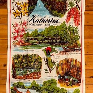 Op de afbeelding: Een vintage theedoek met illustraties van Australische dieren in het wild en bezienswaardigheden uit Katherine, Northern Territory. Het ontwerp bevat een gekapte papegaai, een roodvleugelige papegaai, een zoetwaterkrokodil, een kalkoenstruik, een grevillea, een Gouldian-vink, een kookaburra, een kapokstruik en een uitzicht op Nitmiluk (Katherine Gorge). De tekst "Katherine Northern Territory" is in het midden van de theedoek gedrukt.