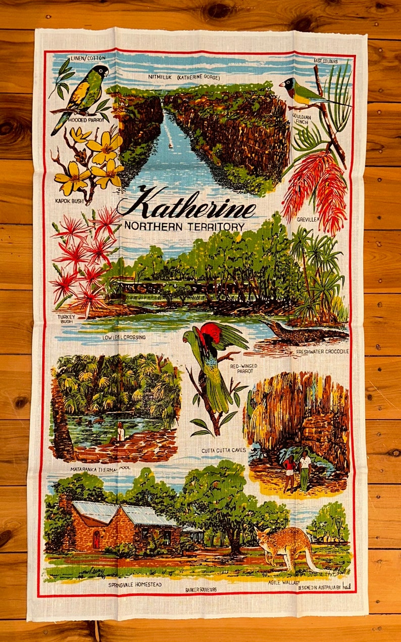 Op de afbeelding: Een vintage theedoek met illustraties van Australische dieren in het wild en bezienswaardigheden uit Katherine, Northern Territory. Het ontwerp bevat een gekapte papegaai, een roodvleugelige papegaai, een zoetwaterkrokodil, een kalkoenstruik, een grevillea, een Gouldian-vink, een kookaburra, een kapokstruik en een uitzicht op Nitmiluk (Katherine Gorge). De tekst "Katherine Northern Territory" is in het midden van de theedoek gedrukt.