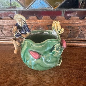 Puede incluir: Un macetero de cerámica verde con forma de hoja con una figura de pescador y una figura de rana en la parte superior. El macetero tiene una flor rosa y hojas verdes.