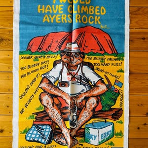 Op de afbeelding: Een kleurrijke theedoek met een cartoonillustratie van een man die op een bankje zit met een koeler en een paar slippers. De tekst op de theedoek luidt "Ik zou Ayers Rock hebben beklommen, maar..." en somt redenen op waarom de man Ayers Rock niet heeft beklommen. De theedoek is gemaakt van linnen en katoen en is kleurvast.