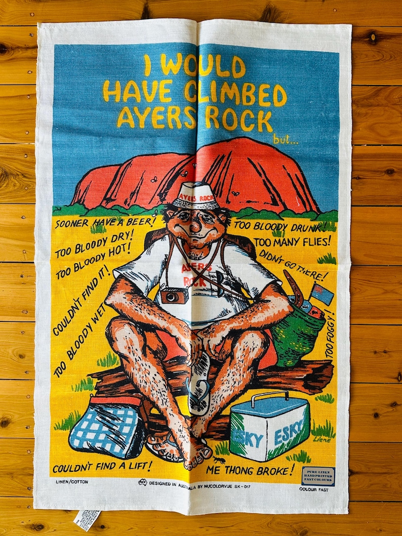 Op de afbeelding: Een kleurrijke theedoek met een cartoonillustratie van een man die op een bankje zit met een koeler en een paar slippers. De tekst op de theedoek luidt "Ik zou Ayers Rock hebben beklommen, maar..." en somt redenen op waarom de man Ayers Rock niet heeft beklommen. De theedoek is gemaakt van linnen en katoen en is kleurvast.