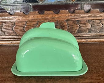 May include: A vintage, mint green butter dish with a lid. The dish has a rectangular base and a rounded, domed lid with a small handle. The butter dish is made of a solid, opaque material and sits on a wooden surface.