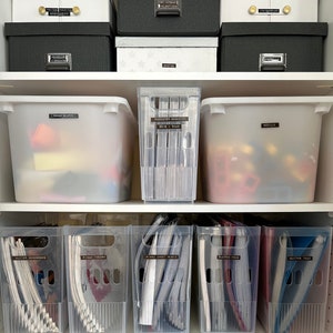 May include: A tidy storage solution with labelled boxes and bins. The top shelf displays white and dark grey boxes with labels such as "Guitar Arrangements" and "Anniversary Valentines." Below are clear bins labelled "Paper Shapes," "Patented Magazine 2010-2023," and "Models."